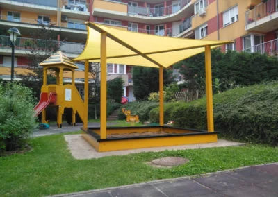 Nagyméretű homokozó sárga napvitorlával lefedve a tűző nap ellen, háttérben lakóházzal.