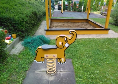 Sárga rugós elefántos játék az előtérben, háttérben egy fedett homokozóval