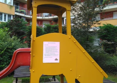 Sárga színű fa játszóvár piros csúszdával és használati útmutatóval egy lakópark udvarán